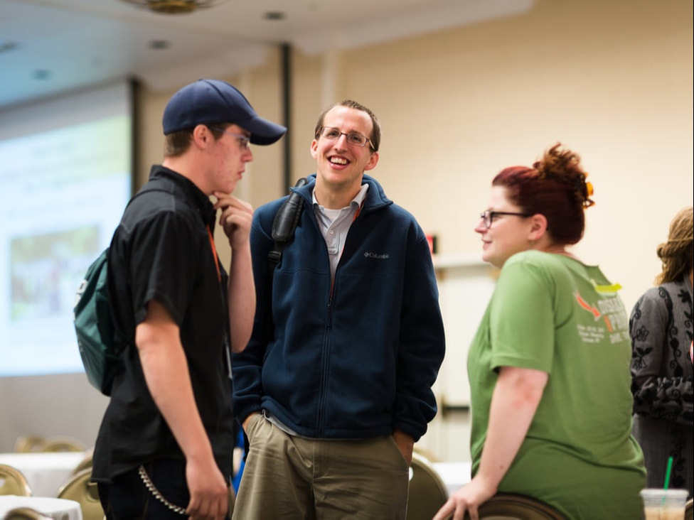 Image of a white woman with red hair in an undo and glasses listening to two young white men. She and on young man are smiling. She is wearing a green tee with Disabled and Proud on the back.